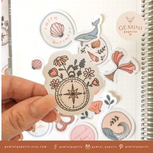 May include: A set of clear stickers with a compass design surrounded by flowers, a whale, seashells, and the text "salty but sweet". The stickers are on a white background with a spiral notebook in the background.