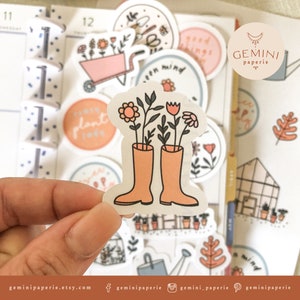 May include: A set of colorful stickers featuring a variety of gardening themes, including flowers, plants, a watering can, a greenhouse, and a pair of rain boots with flowers growing out of them. The stickers are perfect for adding a touch of whimsy to planners, journals, and other surfaces.