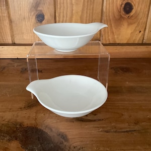 May include: Two white ceramic sauce bowls with a handle-like spout. One bowl is on a clear acrylic stand, and the other is below it. The bowls are on a wooden surface with a wooden background.