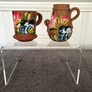 Op de afbeelding: Twee terracotta aardewerkstukken met bloemmotieven geschilderd in roze, blauw, groen en geel. De stukken worden getoond op een heldere acrylstandaard.
