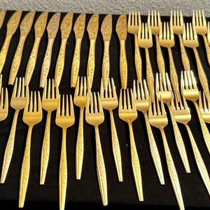 Puede incluir: Una colección de tenedores y cuchillos dorados dispuestos sobre una superficie negra. Los utensilios tienen un diseño texturizado en los mangos. Los tenedores tienen cuatro púas. Los cuchillos tienen punta.