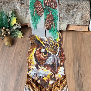 May include: Handmade beaded owl pendant necklace with intricate details. The owl features brown, yellow, and white beads, with a fringe of beads at the bottom. A separate beaded piece depicts pine cones and green needles.