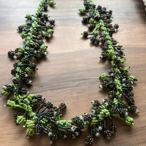Könnte beinhalten: Eine grüne und braune Perlenkette mit einem Blattdesign. Die Kette besteht aus kleinen Perlen und hat ein zartes, filigranes Muster.