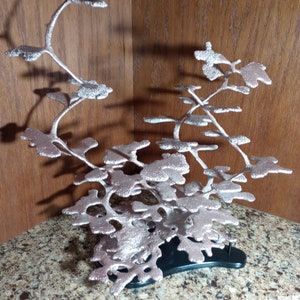 May include: A silver metal sculpture of a tree with a black base. The sculpture is made of many branches and leaves, and it is very detailed. The sculpture is on a granite countertop.