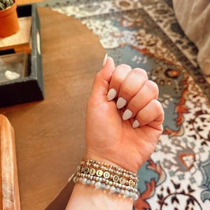 Puede incluir: Una muñeca adornada con múltiples pulseras de cuentas. Las pulseras presentan cuentas doradas y de tonos neutros, con una que deletrea "CHILD OF GOD". La mano tiene esmalte de uñas blanco. El fondo incluye una mesa de madera y una alfombra estampada.