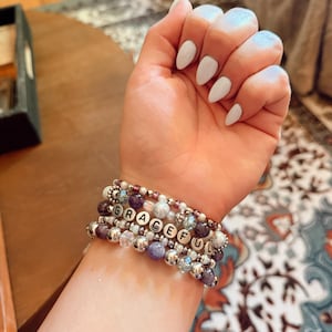 Peut inclure: Plusieurs bracelets de perles avec le mot "GRACEFUL" épelé en perles lettres argentées. Les bracelets combinent des perles violettes, transparentes et argentées. En arrière-plan, une table en bois et un tapis à motifs.