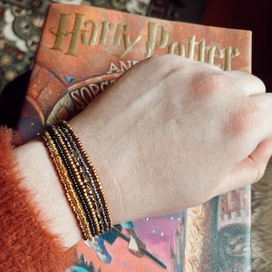 Puede incluir: Una mano con un brazalete de múltiples capas con cuentas doradas y marrones. El brazalete está en la muñeca de una persona, y la mano está apoyada en una copia del libro "Harry Potter y la piedra filosofal".