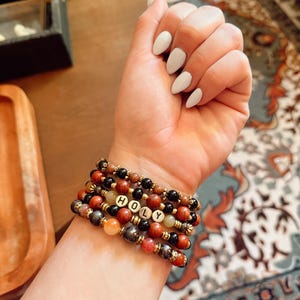 Peut inclure: Plusieurs bracelets de perles rouges, marron, noires et dorées. L'un des bracelets épelle le mot "HOLY" avec des perles de lettres blanches. Les bracelets sont portés sur un poignet avec du vernis à ongles blanc.