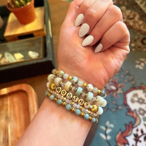 Peut inclure: Plusieurs bracelets de perles bleues, crème et dorées. L'un des bracelets épelle le mot "WORTHY" avec des perles lettres dorées. Les bracelets sont portés sur un poignet avec du vernis à ongles blanc.