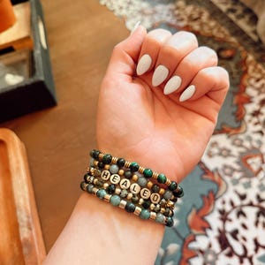 Peut inclure: Plusieurs bracelets de perles vertes, dorées et grises. L'un des bracelets comporte des perles de lettres dorées épelant "HEALED". Les bracelets sont portés sur un poignet avec du vernis à ongles blanc.
