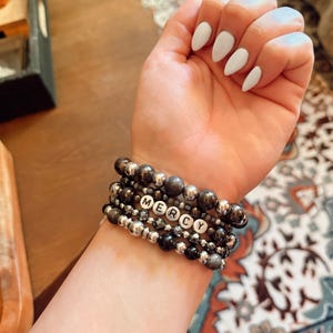 Peut inclure: Plusieurs bracelets de perles grises et argentées. L'un des bracelets comporte des perles de lettres blanches épelant "MERCY". Les bracelets sont portés sur un poignet avec du vernis à ongles blanc.
