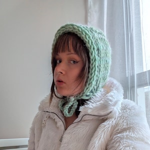 May include: A light mint green hand-knitted bonnet made from thick, chunky yarn. The bonnet features a textured cable-knit pattern and includes two matching knitted ties that fasten under the chin to secure the head covering in place.