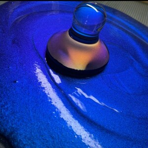 May include: A blue glass bottle stopper with a iridescent finish sits on a blue surface with a white swirl.