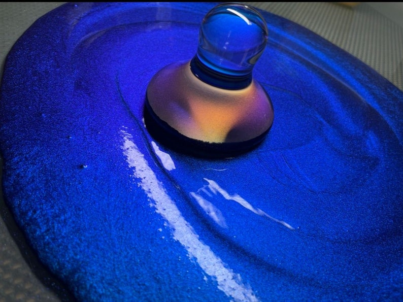 May include: A blue glass bottle stopper with a iridescent finish sits on a blue surface with a white swirl.