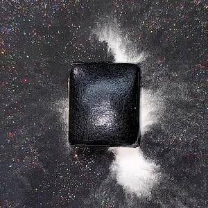 May include: A black square-shaped object with a textured surface, surrounded by white powder on a black background with iridescent glitter.