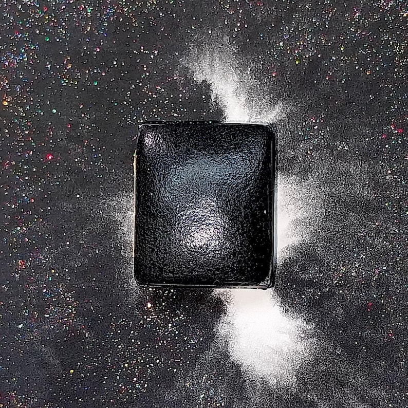 May include: A black square-shaped object with a textured surface, surrounded by white powder on a black background with iridescent glitter.