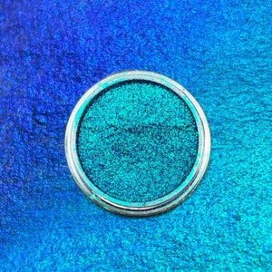 May include: A small round container of teal blue glitter. The glitter is in a clear plastic container with a silver rim.