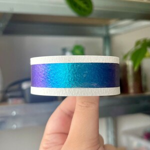 May include: A roll of iridescent washi tape with a white stripe. The tape is blue, green, and purple.