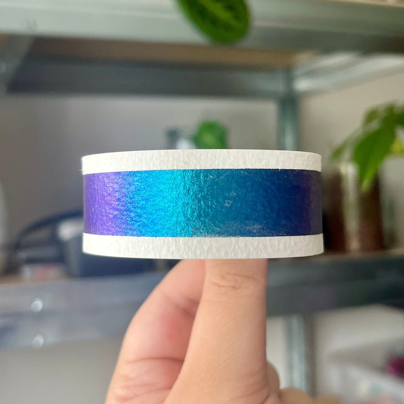 May include: A roll of iridescent washi tape with a white stripe. The tape is blue, green, and purple.