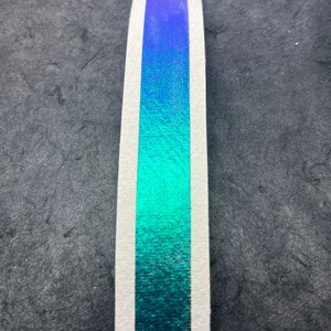 May include: A close-up of a strip of iridescent teal and purple holographic vinyl tape. The tape is on a white background.