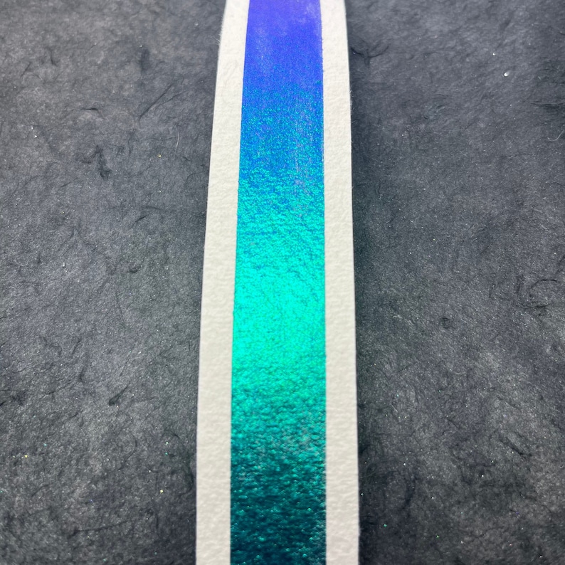 May include: A close-up of a strip of iridescent teal and purple holographic vinyl tape. The tape is on a white background.