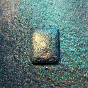Könnte beinhalten: Ein quadratischer Behälter mit schimmerndem Glitzer in den Farben Türkis, Blau und Gold.