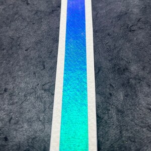 May include: A close-up of a blue and green iridescent tape with a white border. The tape is on a black textured surface.