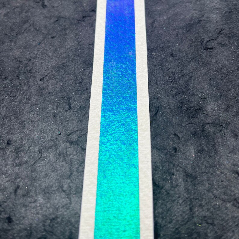 May include: A close-up of a blue and green iridescent tape with a white border. The tape is on a black textured surface.