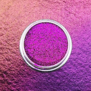May include: A small, round container filled with vibrant, hot pink glitter. The glitter is finely milled and reflects light, creating a sparkling effect. The container has a silver rim and sits on a gradient background of pink and purple.