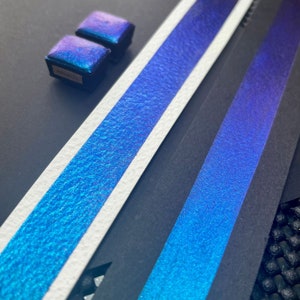May include: Two small, square, iridescent blue watercolor pans on a black surface. The pans are arranged side-by-side, with the top pan slightly overlapping the bottom pan. The pans are surrounded by a white surface with a blue iridescent texture.
