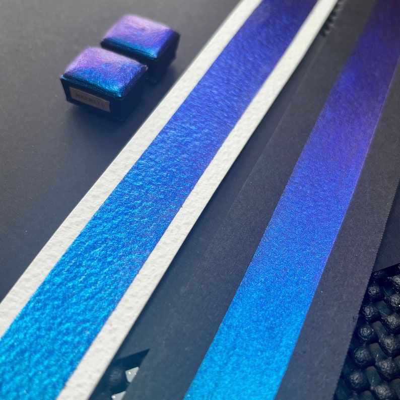 May include: Two small, square, iridescent blue watercolor pans on a black surface. The pans are arranged side-by-side, with the top pan slightly overlapping the bottom pan. The pans are surrounded by a white surface with a blue iridescent texture.
