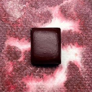 Può includere: Un panetto quadrato di acquerello marrone scuro. La vernice è in un panetto con un bordo bianco. Il panetto è su una superficie strutturata con macchie rosa e bianche.