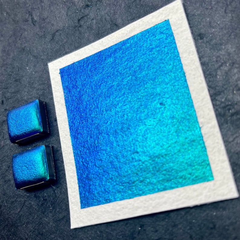 May include: Two small pans of iridescent blue watercolor paint. The paint is a deep blue with a green sheen.