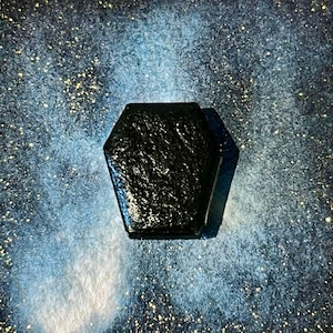 Peut inclure: Un objet noir de forme hexagonale avec une surface texturée. Il est posé sur un fond moucheté bleu et blanc. La forme sombre contraste avec le fond clair et scintillant, créant une image saisissante.