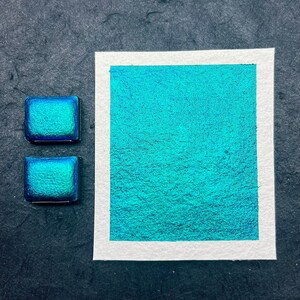 May include: Two small pans of teal blue iridescent watercolor paint. The pans are on a black surface next to a larger sample of the paint on white paper.