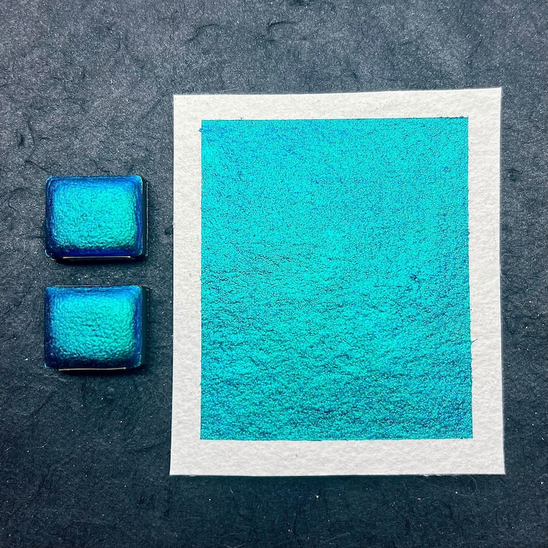 May include: Two small pans of teal blue iridescent watercolor paint. The pans are on a black surface next to a larger sample of the paint on white paper.
