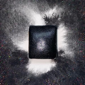 May include: A black square makeup palette with a shiny, reflective surface. The palette is surrounded by white powder, creating a dramatic effect.