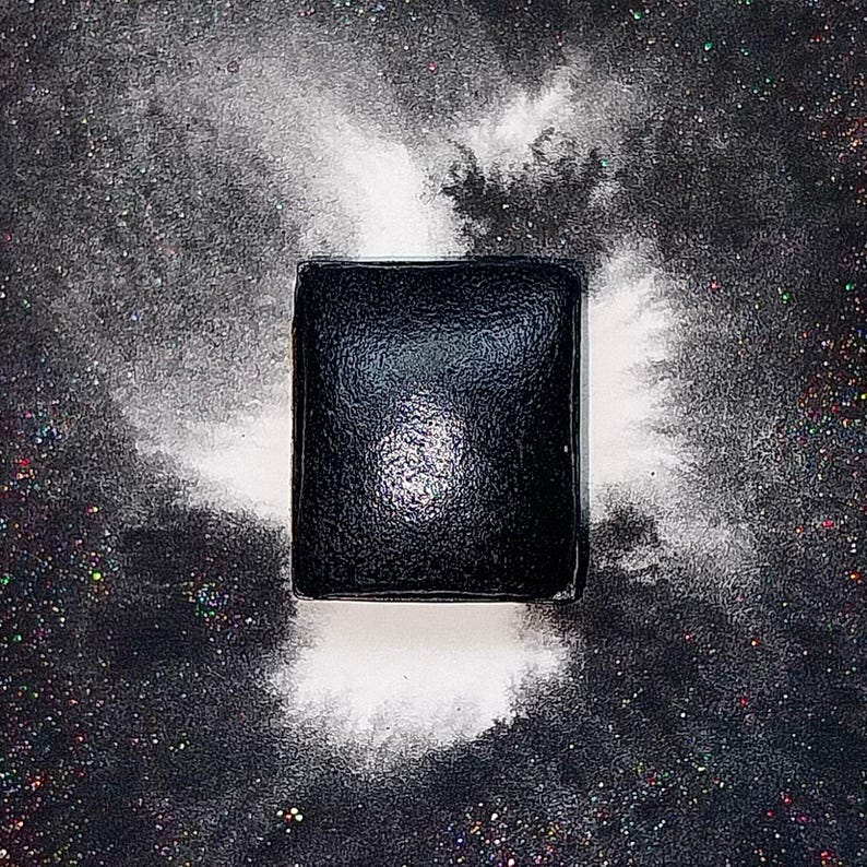 May include: A black square makeup palette with a shiny, reflective surface. The palette is surrounded by white powder, creating a dramatic effect.