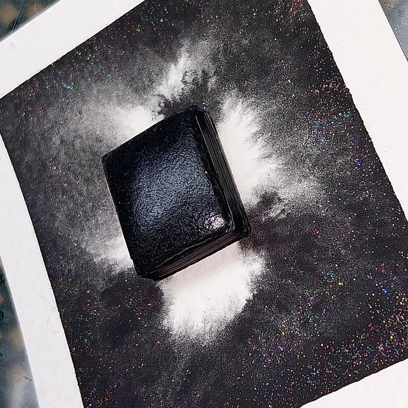 May include: A black square-shaped object with a textured surface is surrounded by white powder on a black background with iridescent glitter.