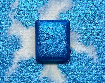 May include: A square, metallic blue paint pan with a textured surface. The pan is surrounded by white paint splatters on a blue background.