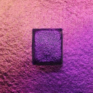 May include: A square purple watercolour pan with a textured surface. The pan is sitting on a pink and purple iridescent background.