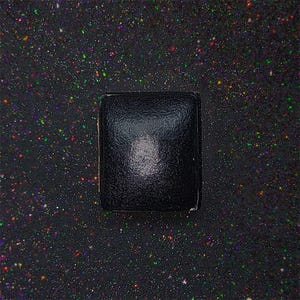 May include: A black square box with a slightly textured surface. The box is sitting on a black background with a scattering of colourful glitter.