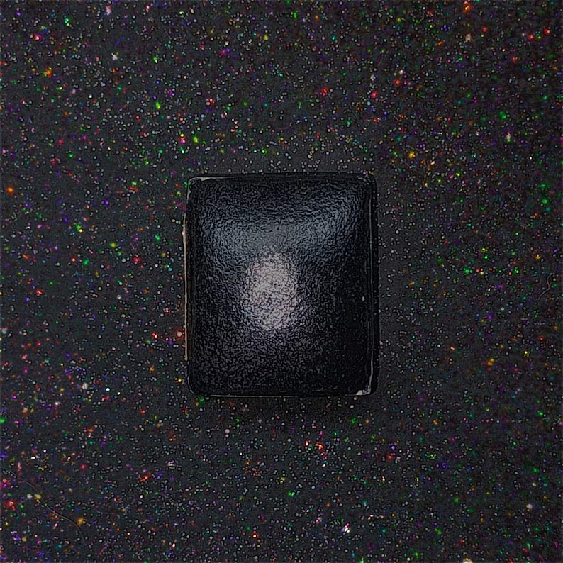 May include: A black square box with a slightly textured surface. The box is sitting on a black background with a scattering of colourful glitter.