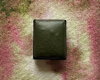 May include: A single, dark green watercolor paint pan in a black plastic container. The pan is full of paint and has a slightly textured surface.