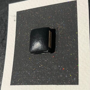 May include: A small black leather box with a gold label that says "CARINE". The box is sitting on a black surface with iridescent glitter.