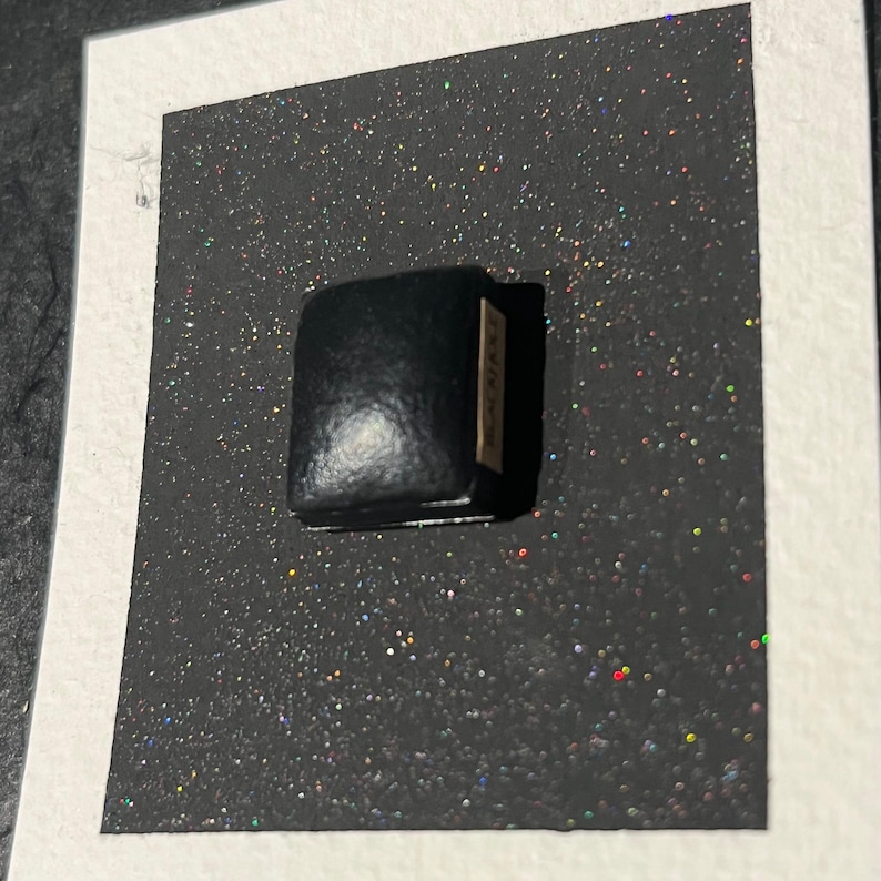 May include: A small black leather box with a gold label that says "CARINE". The box is sitting on a black surface with iridescent glitter.