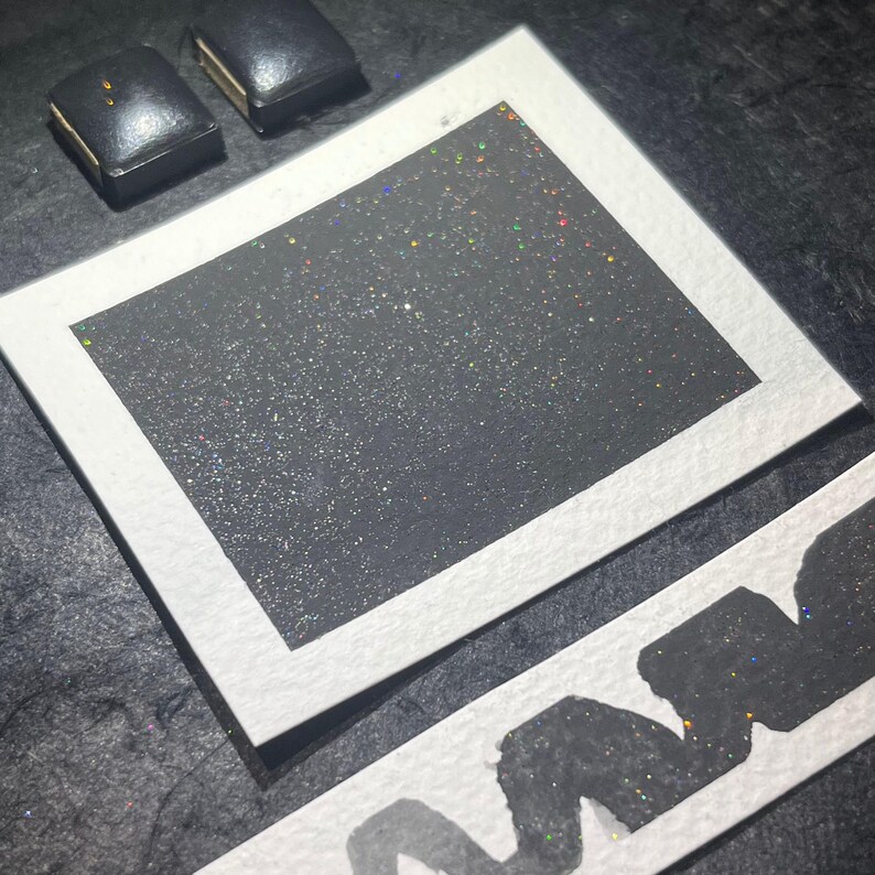 May include: A close-up of a black square with iridescent glitter. The square is framed with a white border and is sitting on a black surface.