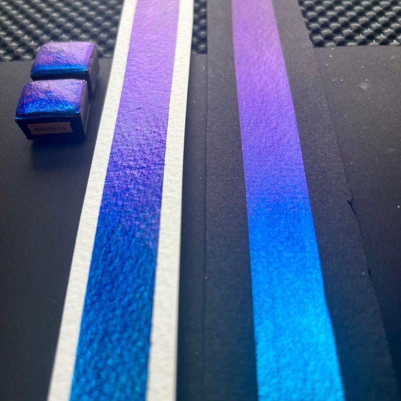 May include: Two small pans of iridescent blue watercolor paint with the word "MEZZANINE" printed on one of the pans. The paint is shown on two white strips of paper, one with a purple to blue gradient and the other with a blue to teal gradient.
