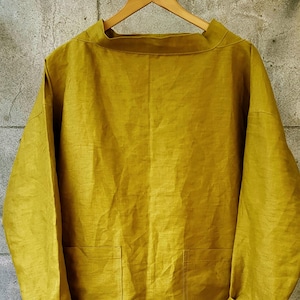 May include: A mustard yellow linen shirt with long sleeves and a boat neck. The shirt has two patch pockets on the front.