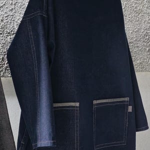 May include: Dark blue denim jacket with two large front pockets and a collar. The jacket has long sleeves and is hanging on a wooden hanger. The stitching is a light color, creating a contrast with the dark denim.